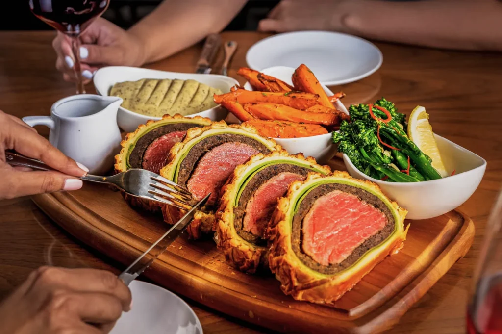 A close-up of a sliced beef Wellington on a wooden board, surrounded by bowls of roasted carrots, broccoli, and sauce. Elegant dining with a glass of wine.