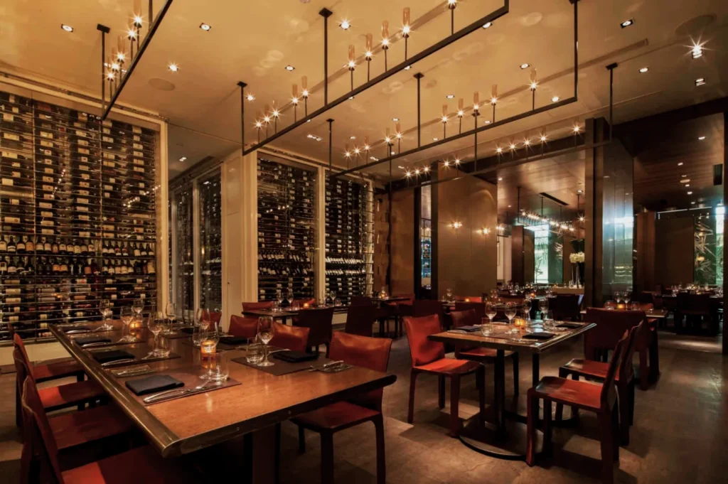 Elegant restaurant interior with warm lighting, brown leather chairs, and neatly set tables. Wine racks line the walls, creating a sophisticated ambiance.