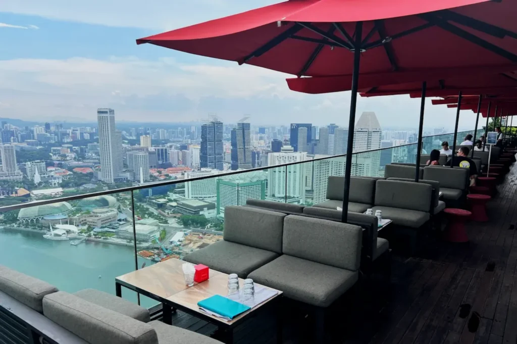 Rooftop lounge with gray sofas and red umbrellas overlooking a city skyline and waterfront. Relaxing atmosphere with a panoramic cityscape view.