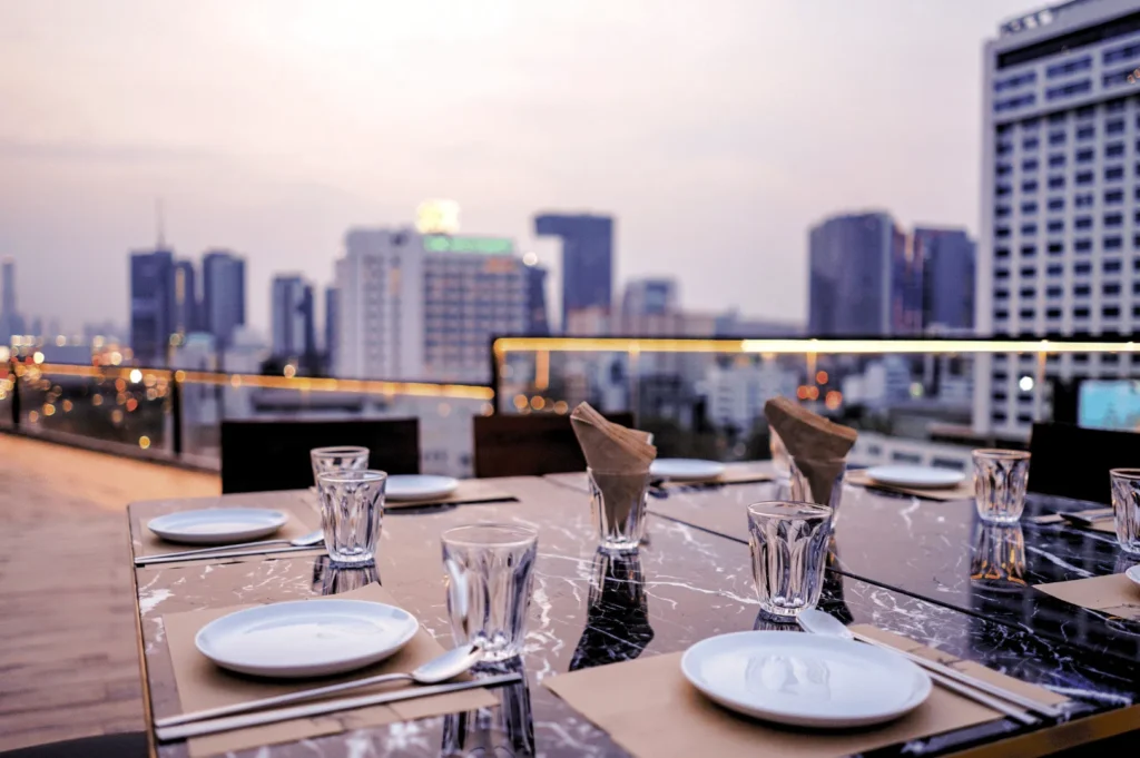 Elegant rooftop dining setup overlooking Singapore’s city skyline at dusk, with marble table settings and warm urban glow—capturing the refined ambiance of high-altitude restaurants that pair panoramic views with sophisticated cuisine.