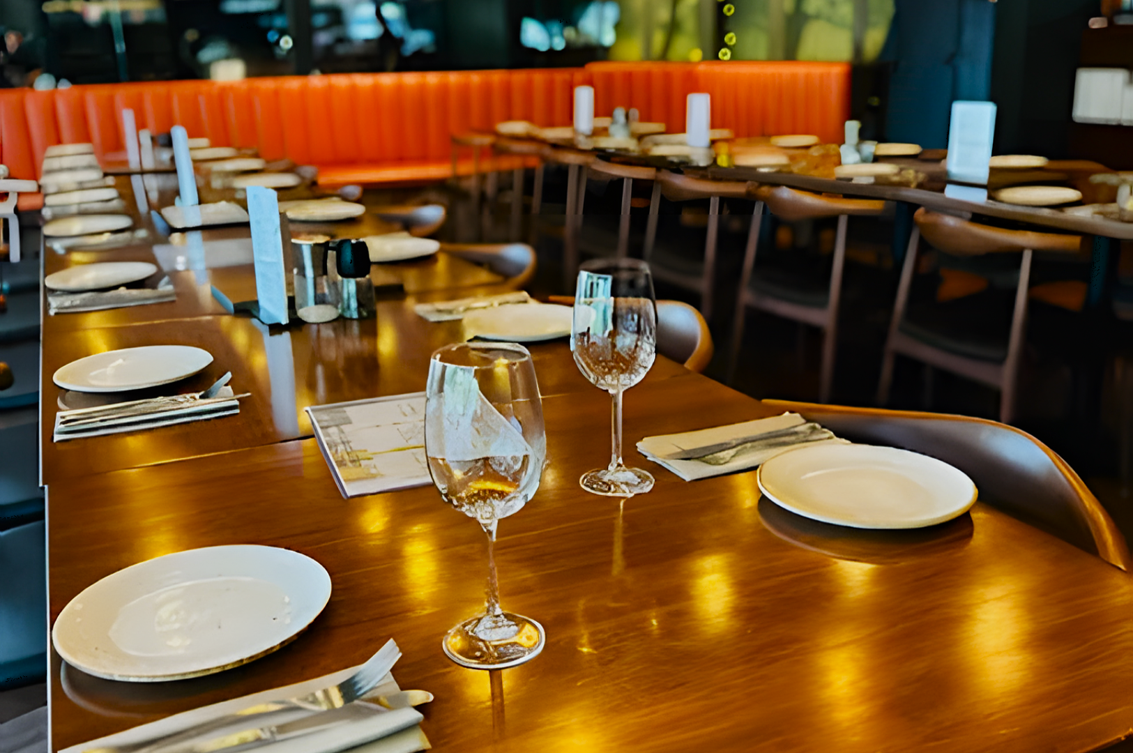 Elegant restaurant interior with neatly set tables, featuring white plates, wine glasses, and cutlery. Orange banquettes and warm lighting create a cozy ambiance.