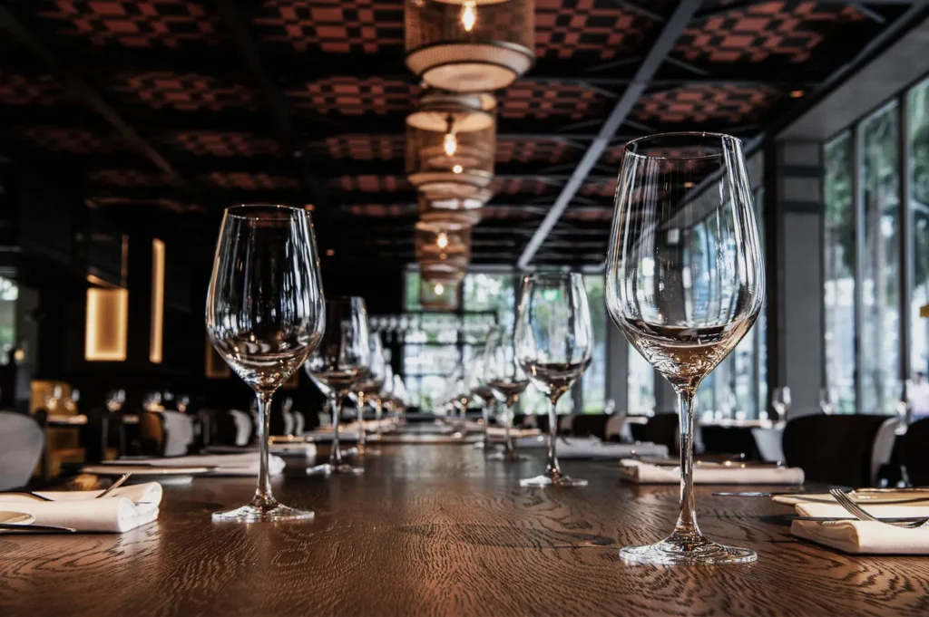 Elegant restaurant interior with large wooden table set with empty wine glasses and rolled napkins. Soft overhead lighting creates a warm, inviting ambiance.