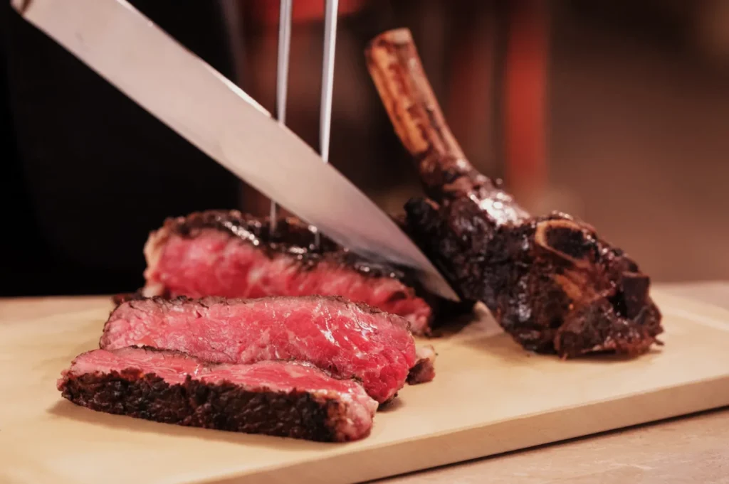 A chef slices a juicy, medium-rare tomahawk steak on a wooden board. The steak is cooked with a charred crust, showcasing a perfectly pink interior.
