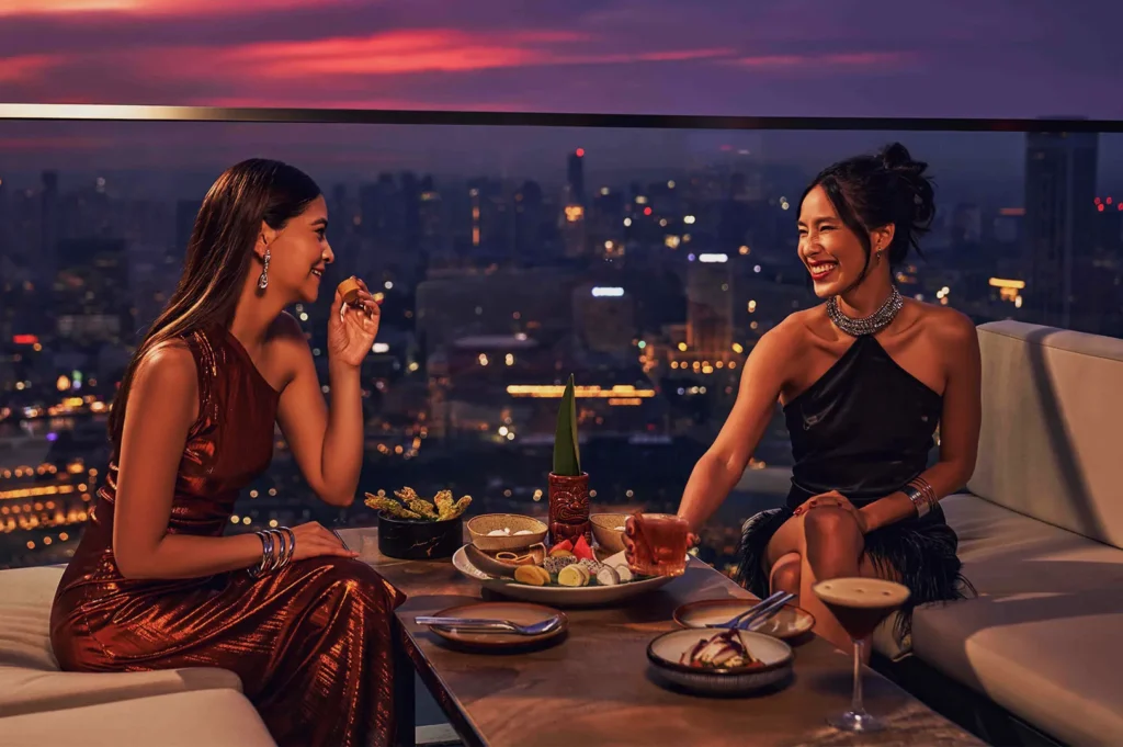 Two women in elegant evening dresses are laughing and enjoying cocktails on a rooftop lounge at sunset, overlooking a city skyline. Plates of appetizers are on the table.