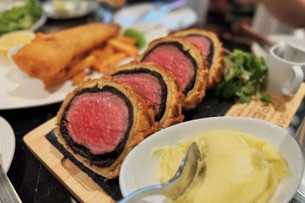 Close-up of sliced beef Wellington with golden pastry on a wooden board, accompanied by mashed potatoes. A background of fish and chips adds depth.