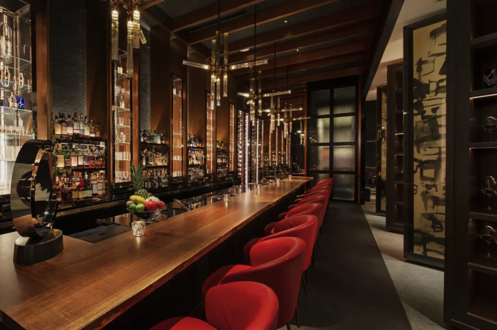 A sleek, modern bar interior with warm lighting, featuring a long wooden counter lined with vibrant red chairs. Elegant pendant lights hang above, adding to the cozy ambiance.