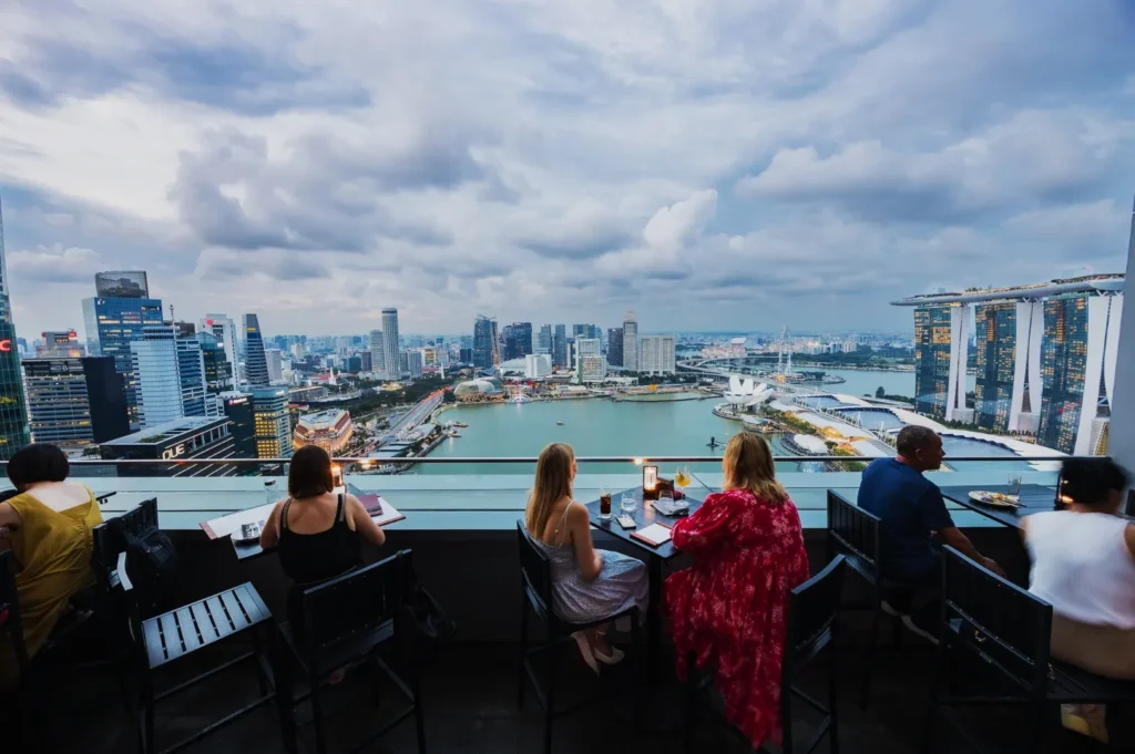 People seated on a rooftop bar enjoy a stunning view of Singapore's skyline and Marina Bay Sands under a cloudy sky, creating a serene atmosphere.
