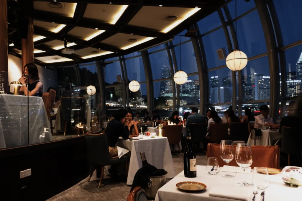 Upscale restaurant interior with floor-to-ceiling windows framing Singapore’s glittering skyline at night; warm lighting, elegant furnishings, and candlelit tables evoke a luxurious dining experience where city views meet refined hospitality.