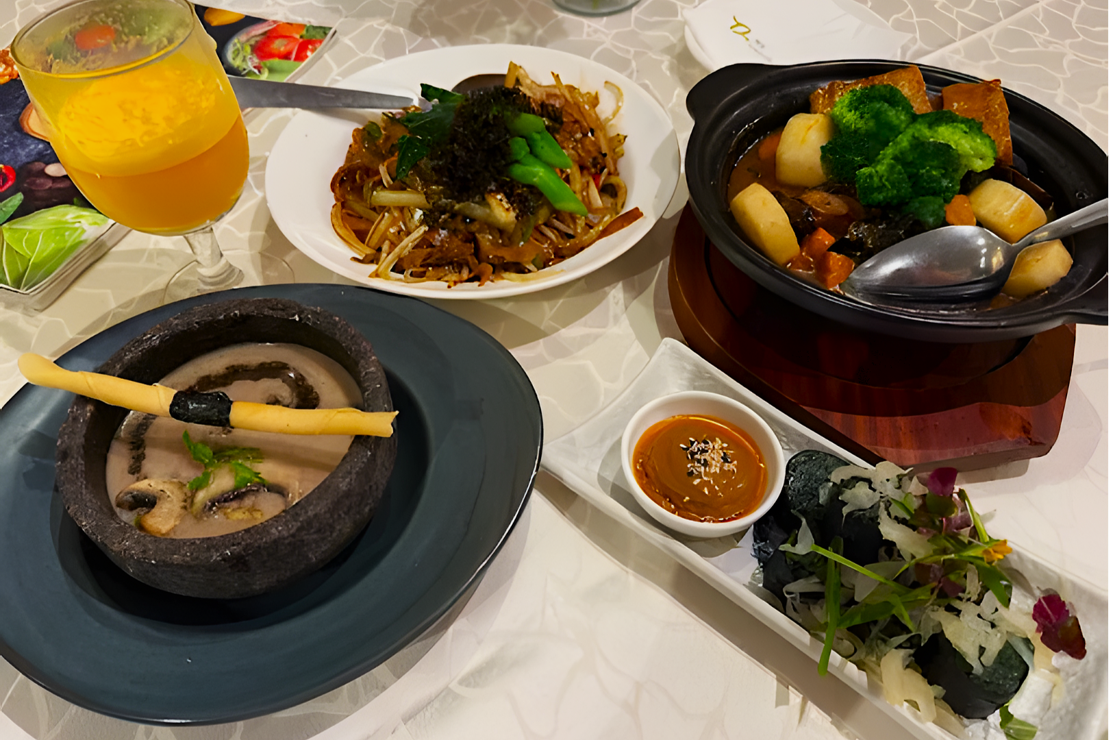 A vibrant meal on a table features a slate bowl of creamy soup with a breadstick, a plate of stir-fried noodles with green garnish, a hot pot with broccoli and mushrooms, sushi rolls on a white tray with dipping sauce, and a glass of orange juice. The setting conveys a sense of gourmet dining.