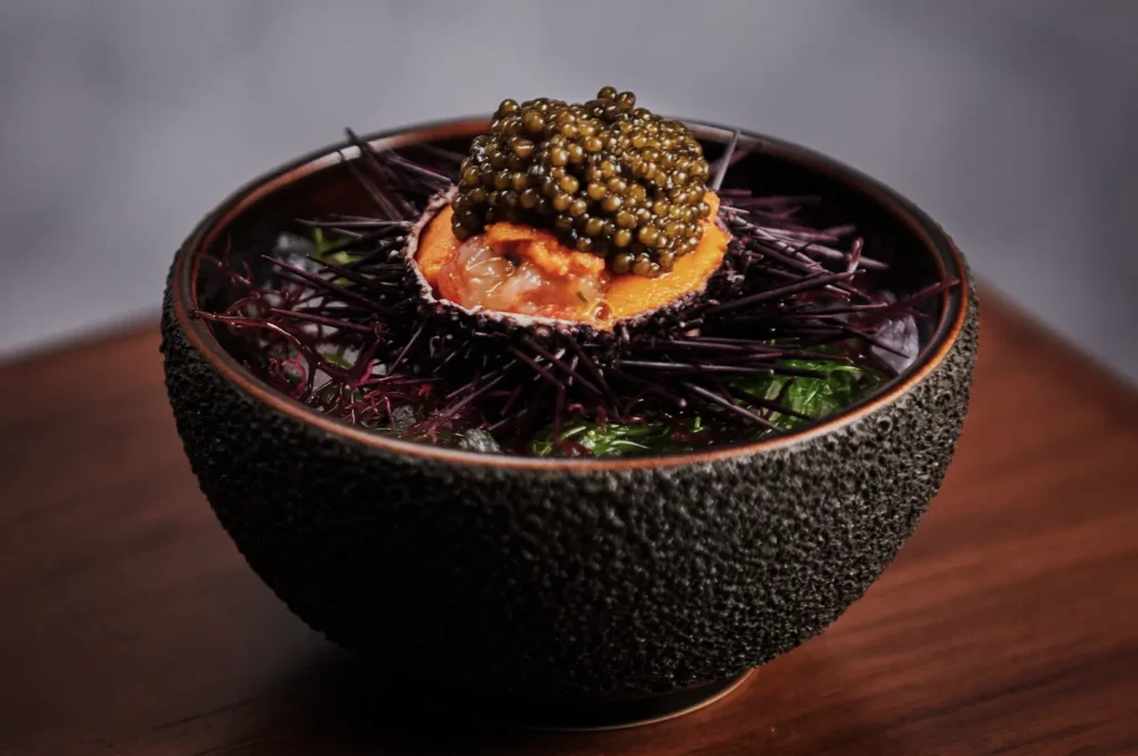 A textured black bowl holds a sea urchin filled with bright orange roe and topped with caviar. The dish is garnished with seaweed, creating a luxurious and elegant presentation.