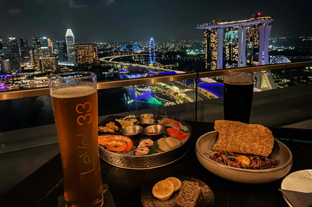 Rooftop dining scene with a city view at night. A seafood platter and two beers, with illuminated skyscrapers and a lit Ferris wheel in the background.