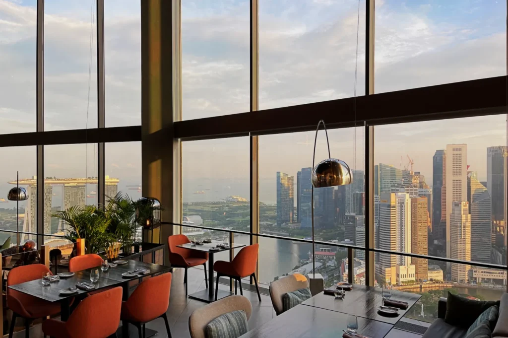 Spacious, modern restaurant with orange chairs and large windows showing a cityscape with high-rise buildings and a distant waterfront at sunset.