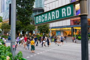 Crowd crossing a busy street at Orchard Road, Singapore. Visible are diverse pedestrians, modern buildings, and a prominent street sign, conveying urban vibrancy.