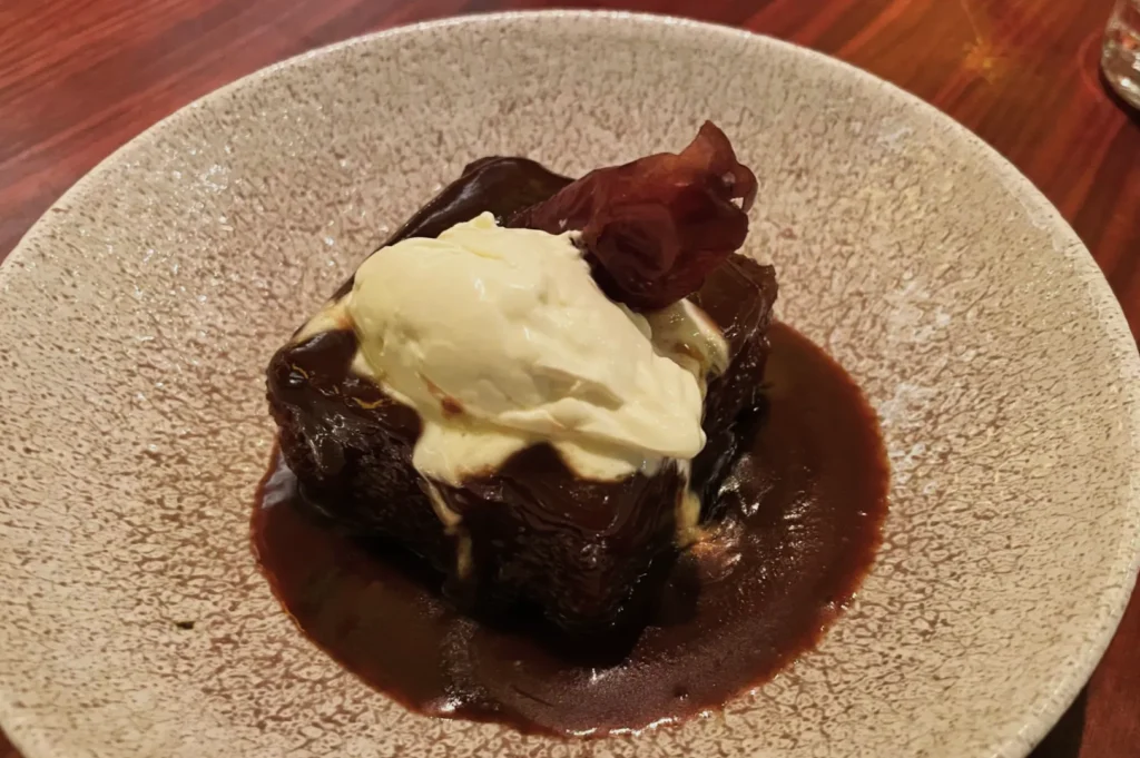 A rich dessert on a speckled plate features a dark chocolate cake topped with creamy vanilla ice cream, garnished with a piece of candied fruit.
