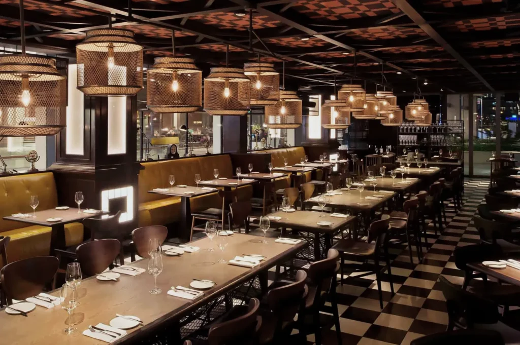 Stylish restaurant interior with checkered floor, warm lighting from woven pendant lamps, and neatly set tables with wine glasses and napkins.
