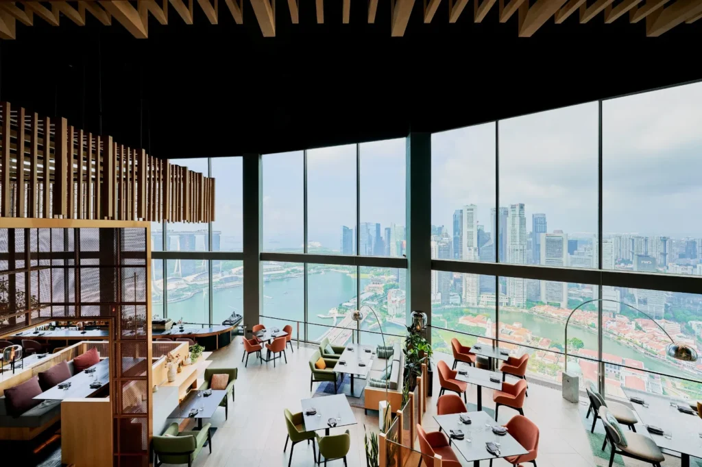 Modern restaurant interior with large windows revealing a city skyline and waterfront. Elegant wood accents and colorful chairs create a sophisticated ambiance.