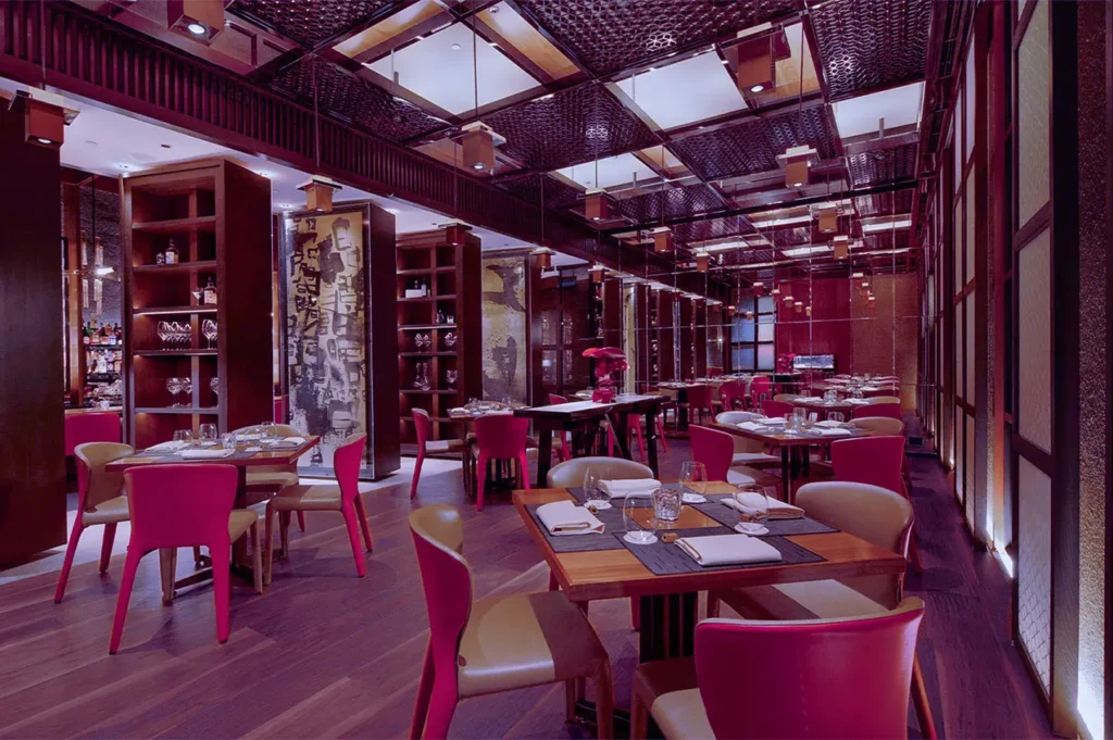 Elegant restaurant interior with wooden tables, red chairs, and soft lighting. The ambience is warm, sophisticated, and inviting, with art-filled walls.