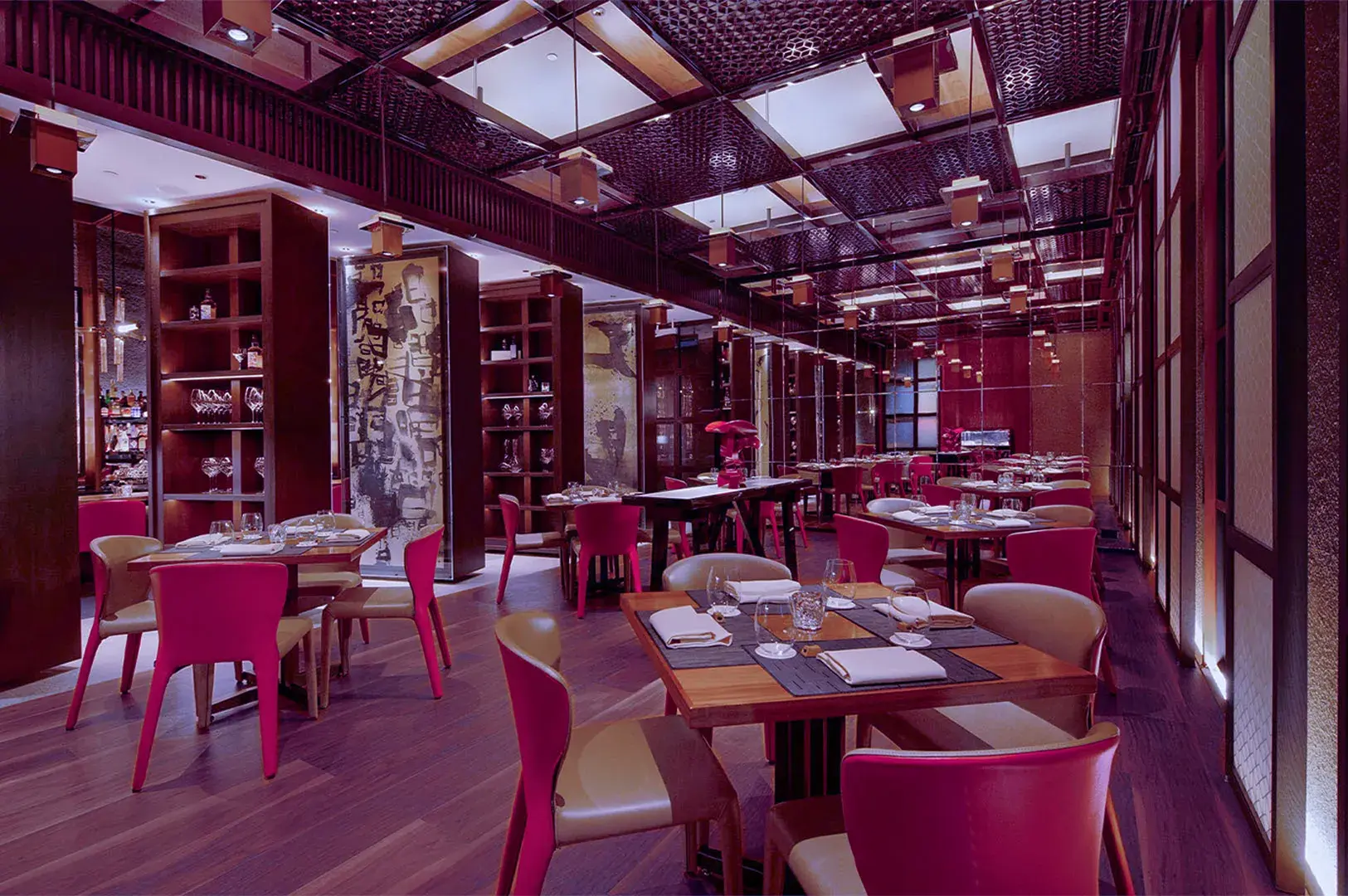 Elegant restaurant interior with wooden tables, red chairs, and soft lighting. The ambience is warm, sophisticated, and inviting, with art-filled walls.