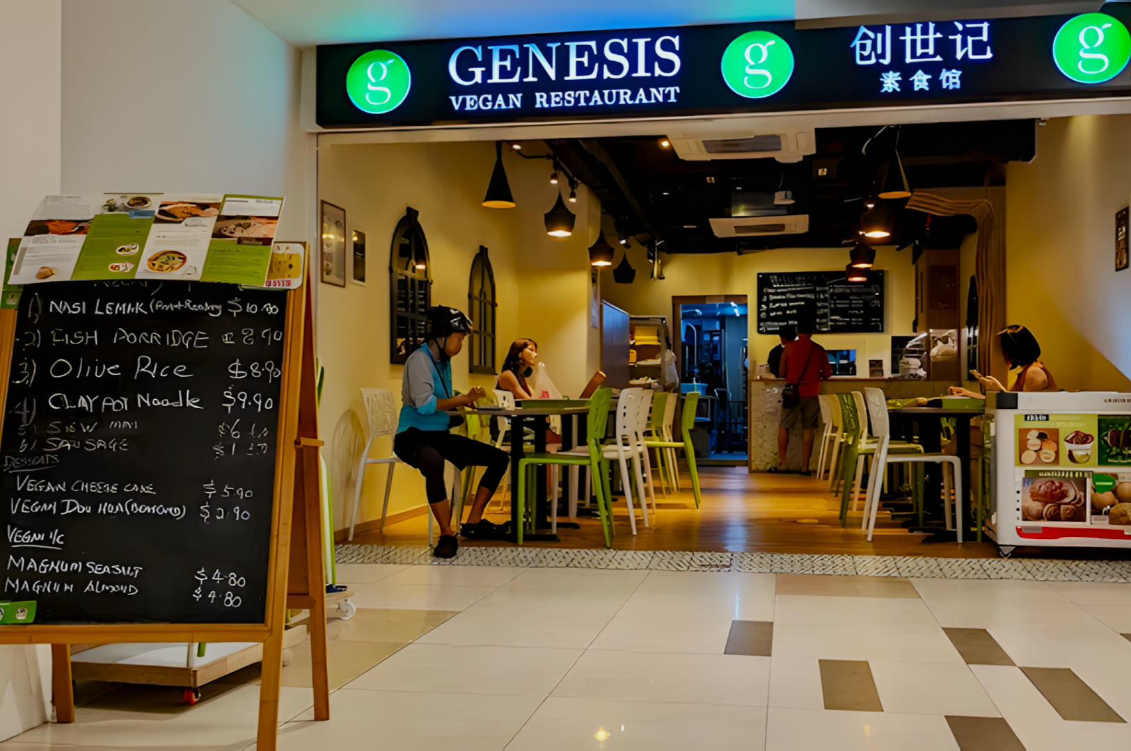 Cozy vegan restaurant entrance with a blackboard menu listing dishes like Nasi Lemak and Olive Rice. Patrons sit at tables, creating a relaxed atmosphere.