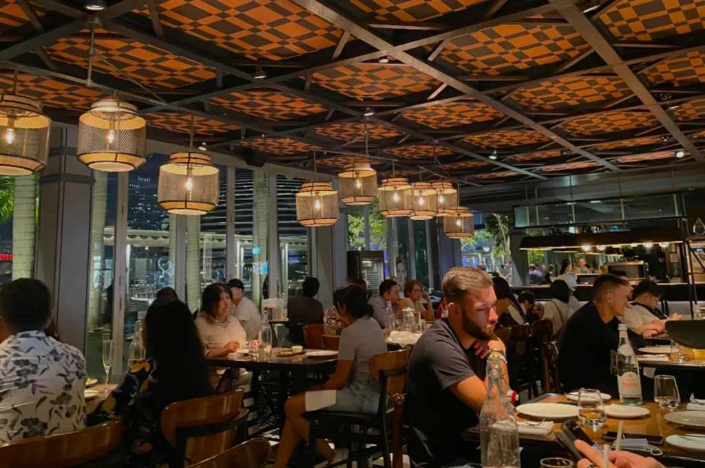 Modern restaurant interior with dim lighting and woven pendant lamps. Tables are filled with diners engaged in conversation, creating a lively atmosphere.