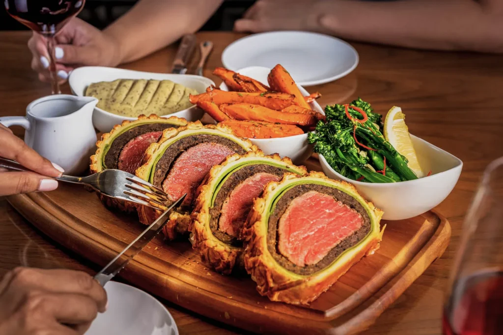 A wooden platter with sliced Beef Wellington, bright pink and wrapped in golden pastry. Sides include roasted carrots, broccoli, and sauces. Cozy dining setting.