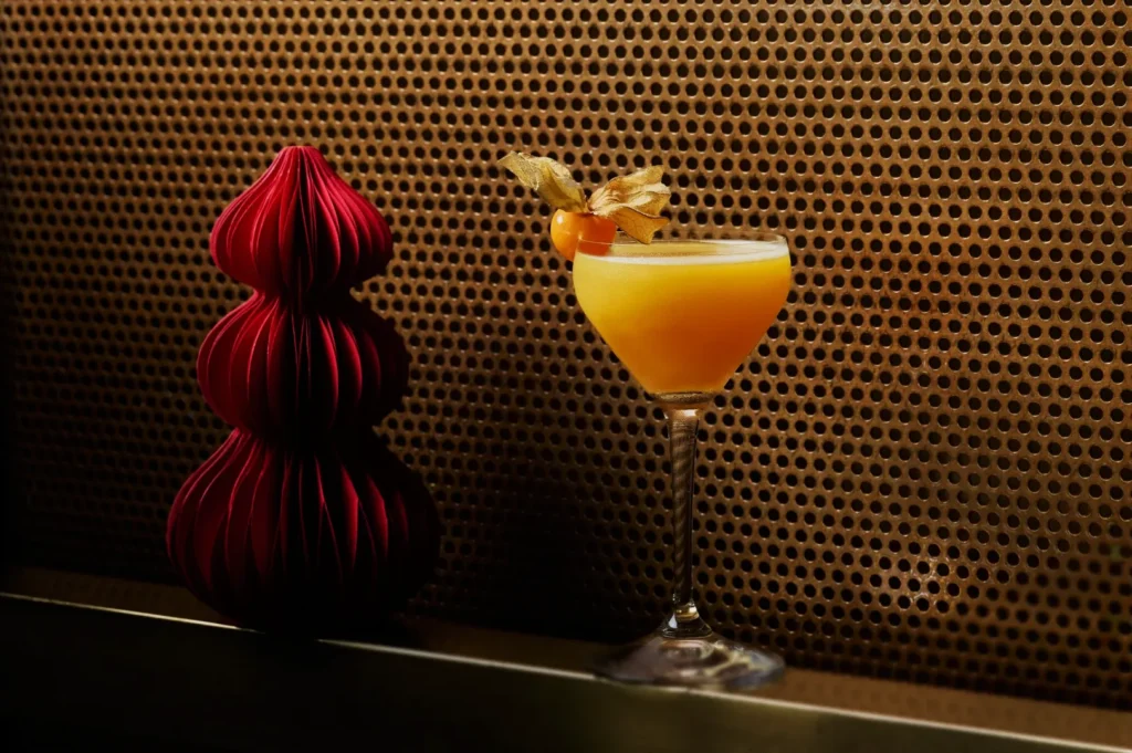 A vibrant orange cocktail garnished with a golden berry in a sleek glass sits beside a red, textured ornament. The background features a perforated metal pattern.