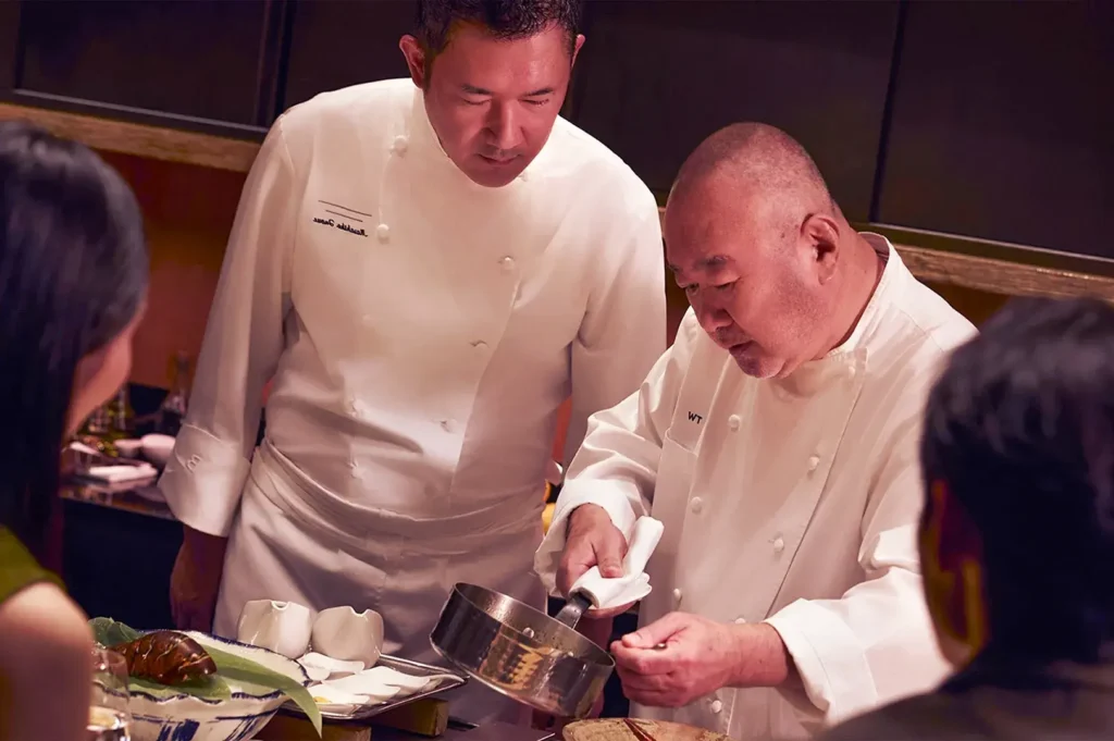 Two chefs in white uniforms collaborate in a warm-toned kitchen. One chef attentively watches as the other prepares a dish, conveying focus and expertise.