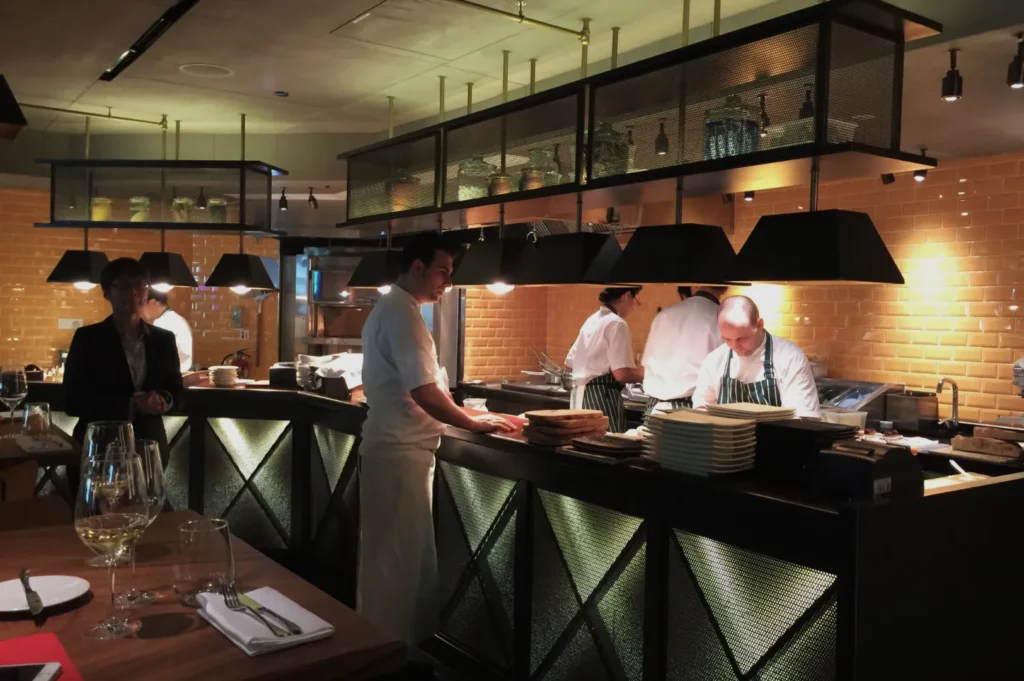 Chefs in a modern, warmly-lit kitchen prepare dishes behind a counter with stacked plates. A customer stands nearby. Wine glasses set the dining ambiance.