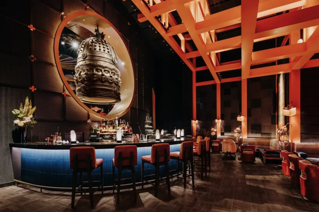 A dimly lit, elegant bar with red chairs and blue accents. A large, ornate sculpture and warm lighting create a sophisticated, inviting atmosphere.