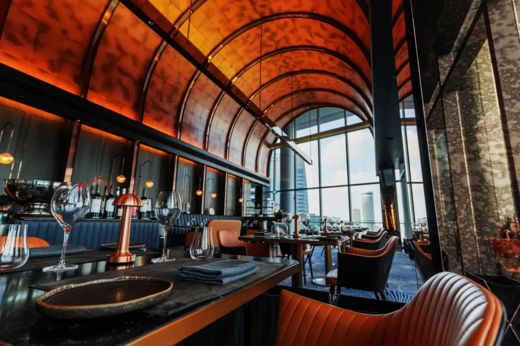Luxurious restaurant interior with orange leather chairs, elegant table settings, and a striking arched ceiling. Large windows offer a cityscape view.