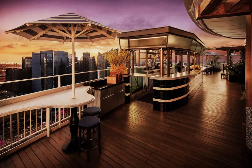 Rooftop bar with wooden flooring and modern furnishings overlooks city skyscrapers at sunset. Cozy seating and warm lighting create an inviting atmosphere.