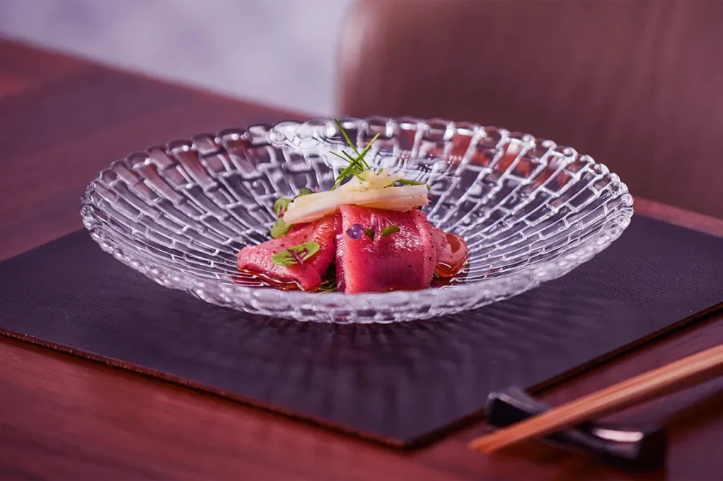 A delicate serving of tuna sashimi garnished with herbs and onions on a textured glass plate. The setting is elegant, with a serene and refined tone.
