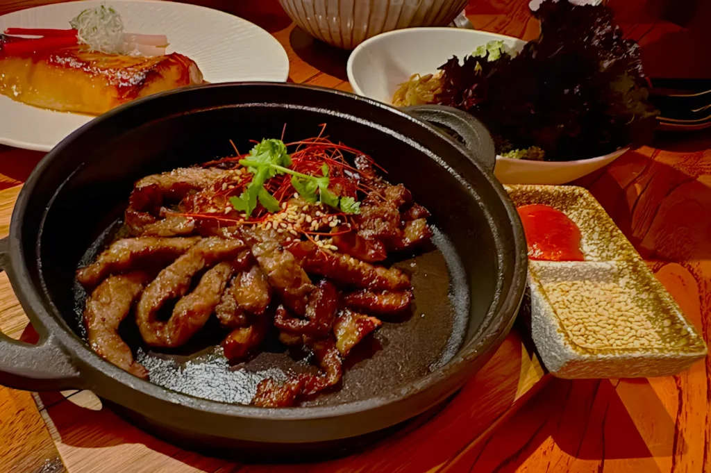 Grilled beef slices in a black skillet garnished with herbs and sesame seeds; a plate with caramel dessert in the background, creating a rich, savory scene.