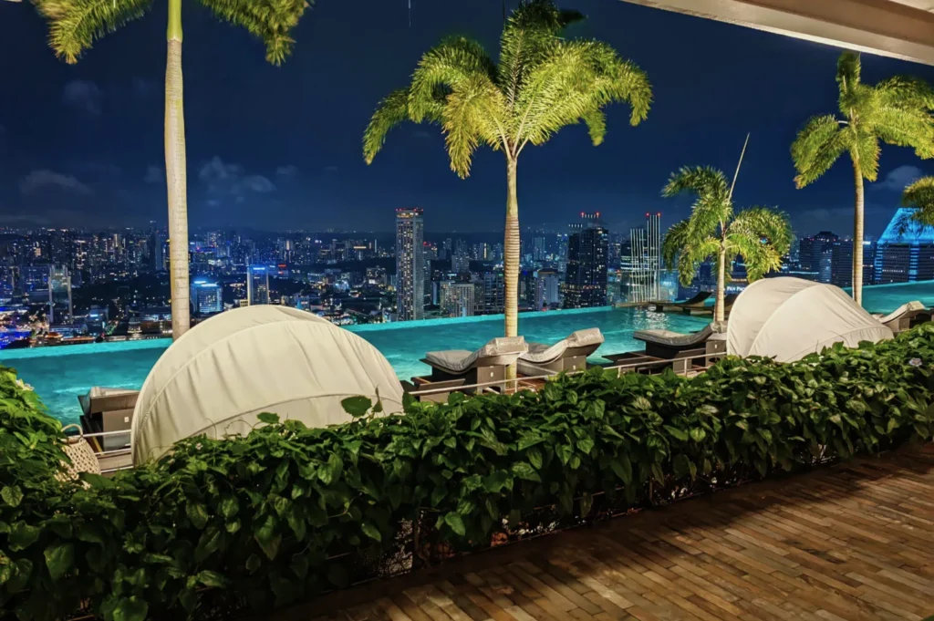 Nighttime cityscape with a rooftop infinity pool and palm trees, overlooking a brightly lit skyline. Cozy lounge areas create a serene and luxurious atmosphere.