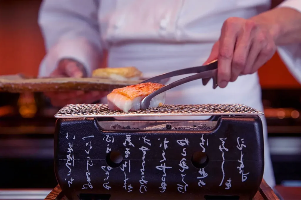 Chef grilling fish on a small, decorative hibachi grill with tongs. The setting is intimate, showcasing precision and culinary artistry.