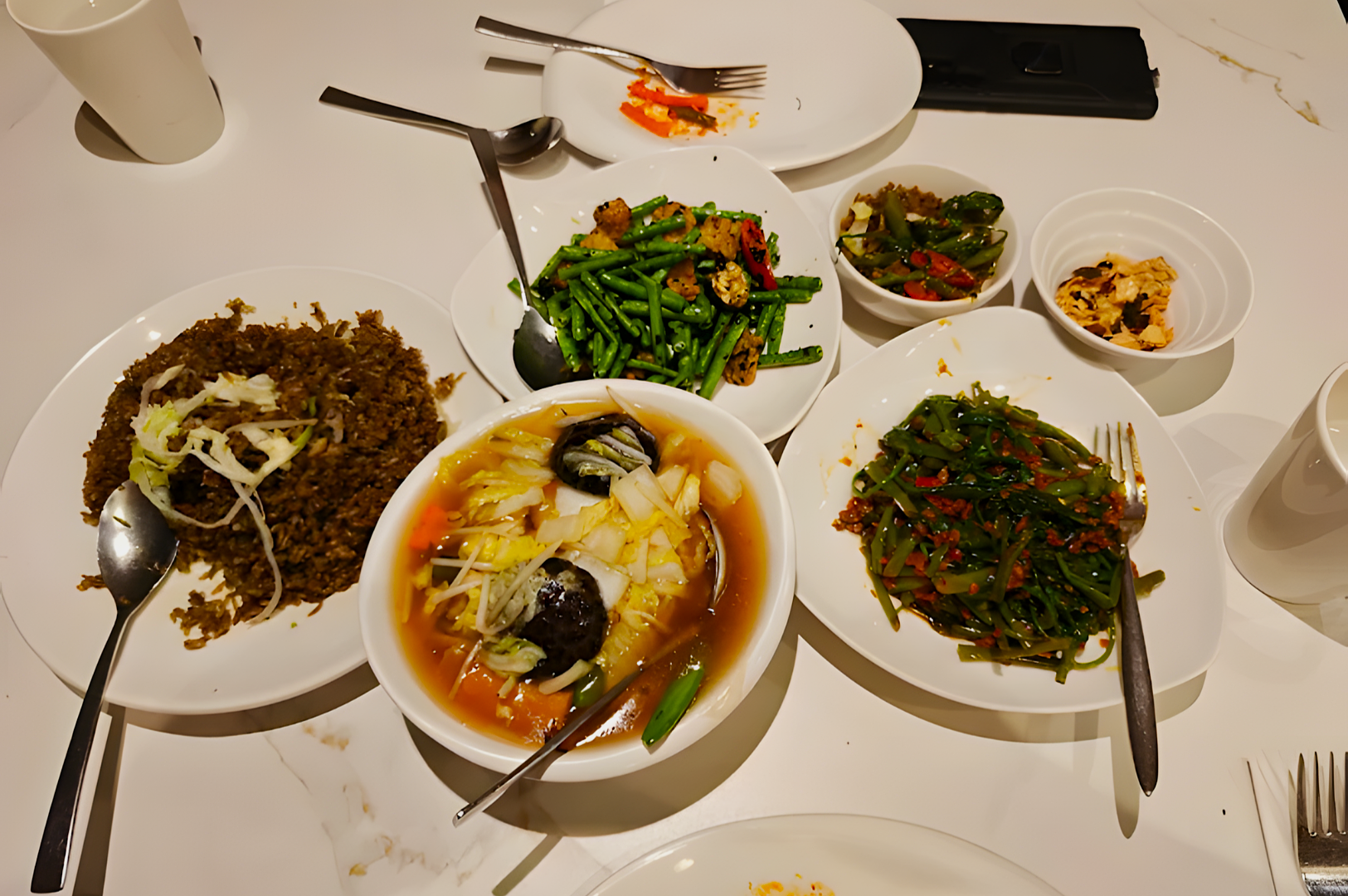 A white table with Asian dishes: fried rice, soup with greens, sautéed vegetables, spicy greens, and dipping sauces. Plates and cutlery suggest a shared meal.
