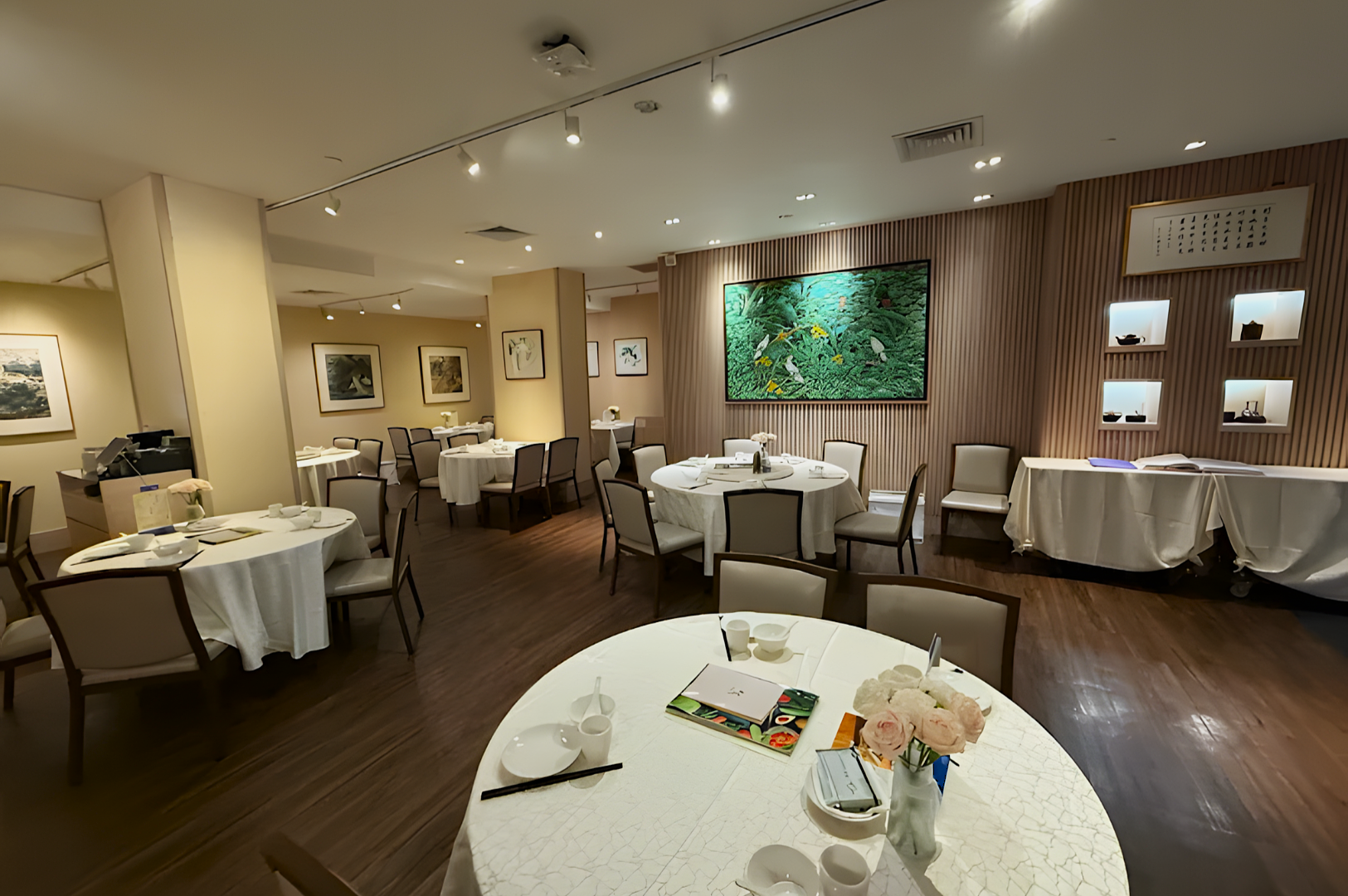 Elegant restaurant interior with round, white-clothed tables and chairs on wooden flooring. Artwork adorns the walls, and soft lighting creates a calm ambiance.