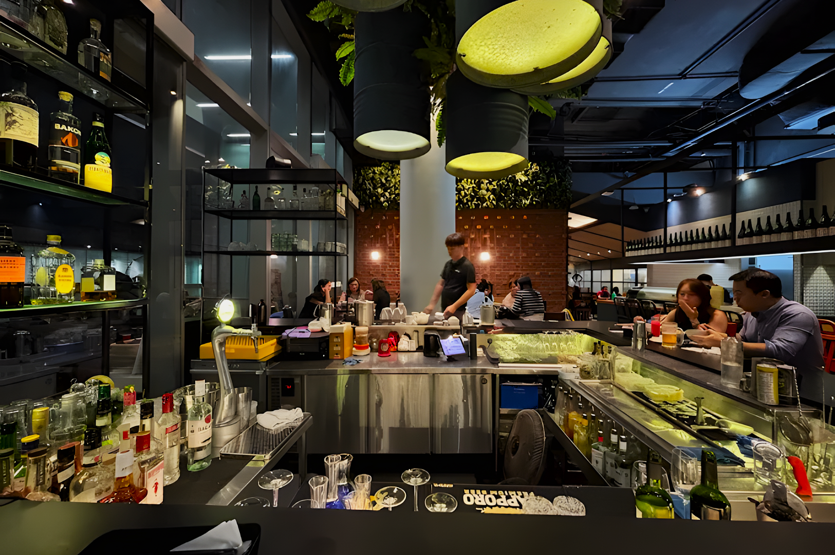 A modern bar with hanging lights, bottles displayed, and patrons seated at a counter. The bartender prepares drinks in a lively, warm setting.
