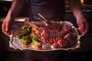 A person holds a silver tray with a large grilled steak, sliced for serving, garnished with herbs. The setting is warm and inviting, suggesting fine dining.