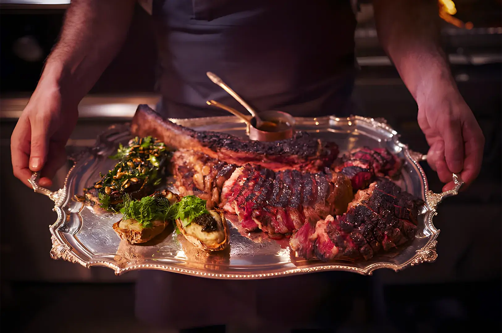 A person holds a silver tray with a large grilled steak, sliced for serving, garnished with herbs. The setting is warm and inviting, suggesting fine dining.