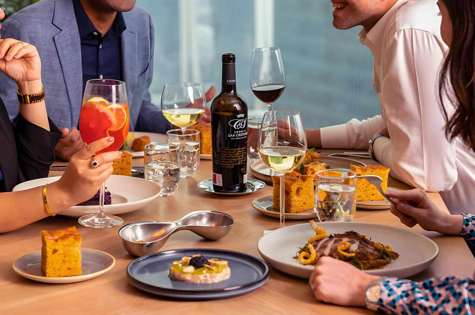 A group of people enjoy a meal at a wooden table, sharing wine and dishes. The scene is lively, with bright food and laughter, creating a joyful atmosphere.