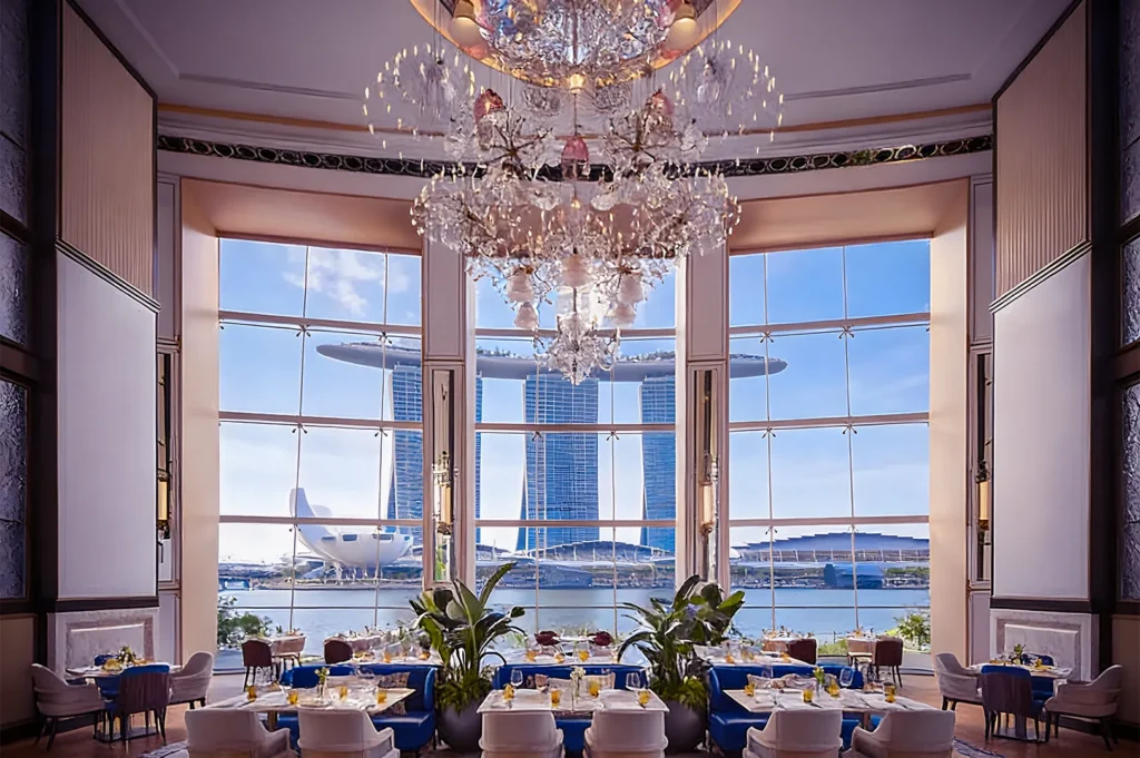 Luxurious dining room with elegant chandelier and large windows showcasing Marina Bay Sands. Blue sky, opulent setting, and a grand ambiance.