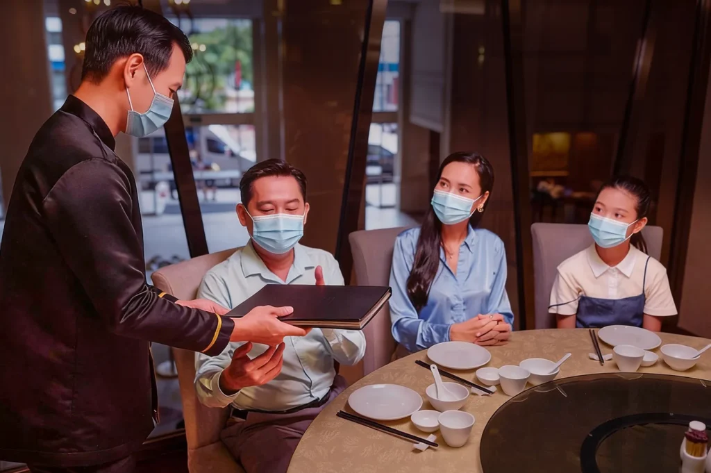 A family in masks sits at a restaurant table with empty dishes. A server, also masked, hands a menu to the father, creating a focused and respectful atmosphere.
