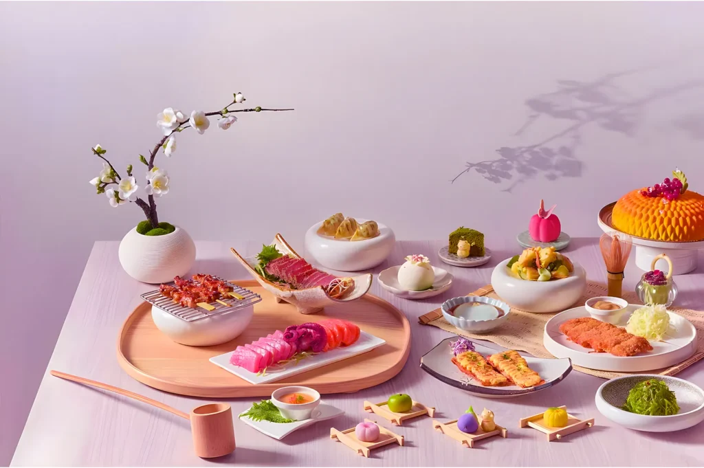 Elegant Japanese meal on a light pink table with sushi, sashimi, grilled meats, and colorful mochi. White flowers in a vase add a serene touch.