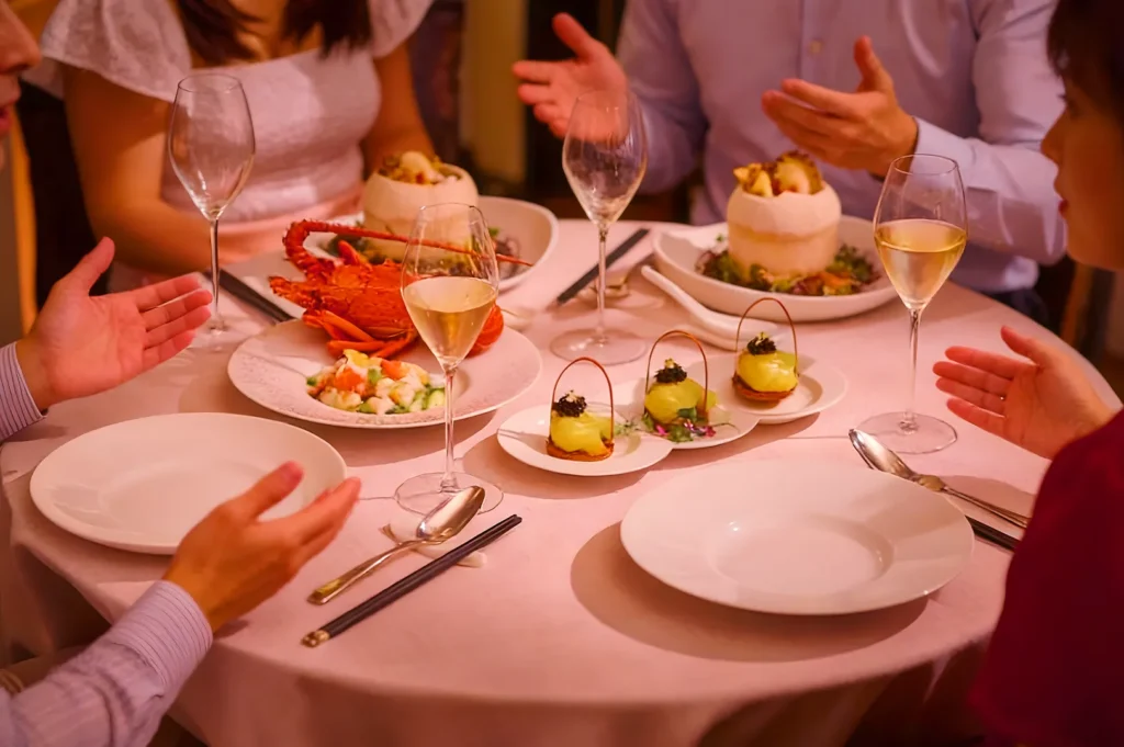 A table with four people half-visible, engaged in conversation. Dishes include lobster, seafood salad, and appetizers, set alongside wine glasses on a pink tablecloth.