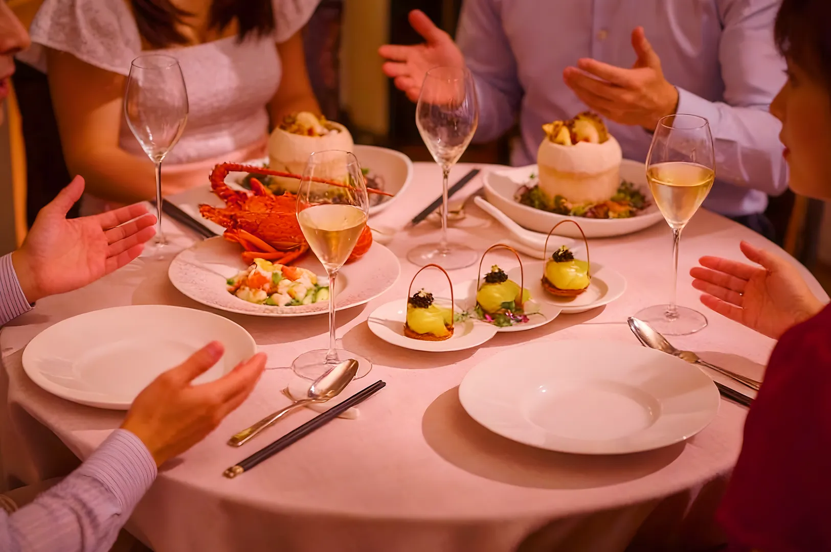 A table with four people half-visible, engaged in conversation. Dishes include lobster, seafood salad, and appetizers, set alongside wine glasses on a pink tablecloth.