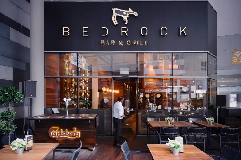 Exterior view of Bedrock Bar & Grill entrance featuring a modern glass facade, outdoor wooden seating, and a server entering the restaurant.