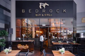 Exterior view of Bedrock Bar & Grill entrance featuring a modern glass facade, outdoor wooden seating, and a server entering the restaurant.