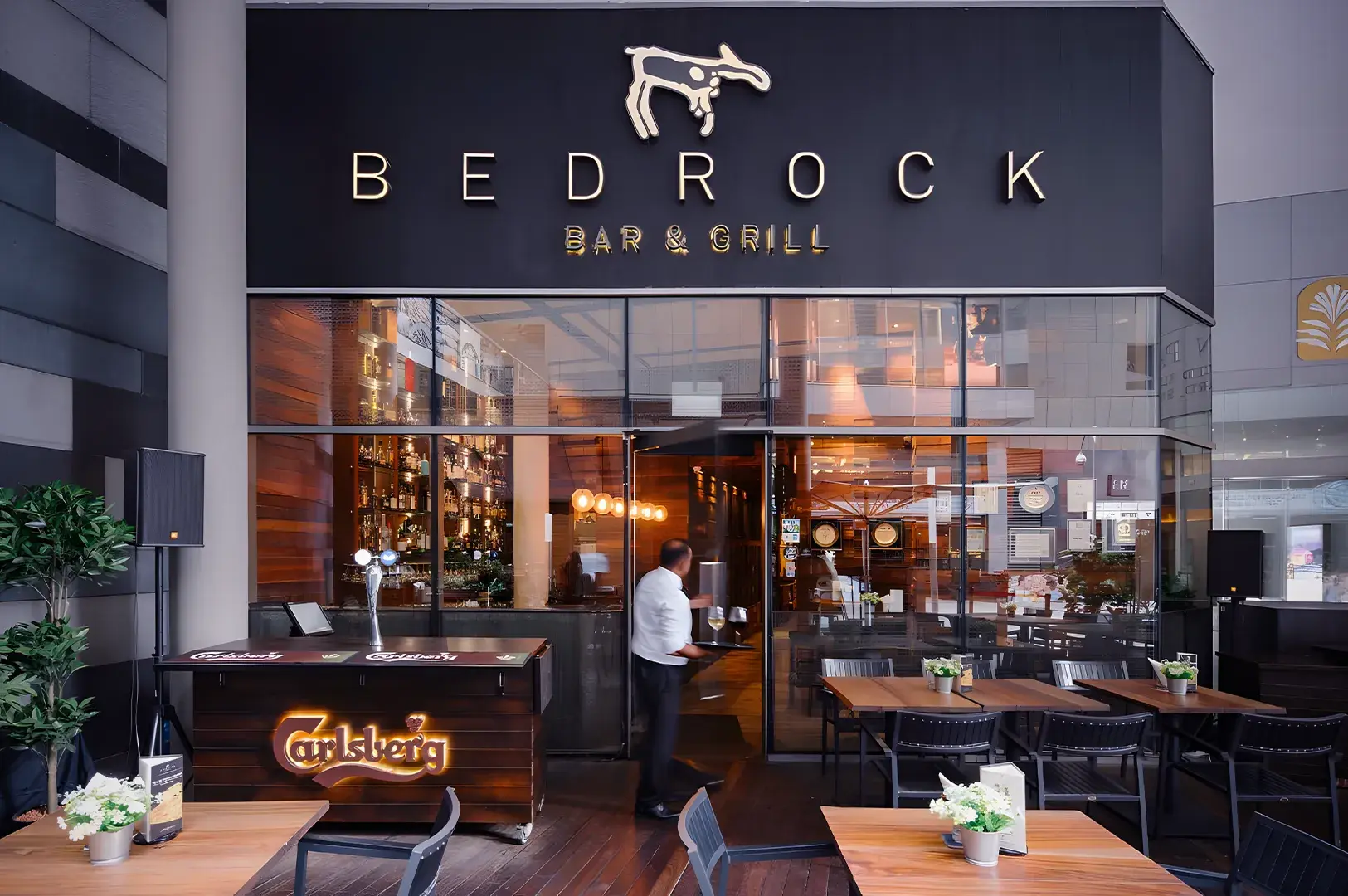Exterior view of Bedrock Bar & Grill entrance featuring a modern glass facade, outdoor wooden seating, and a server entering the restaurant.