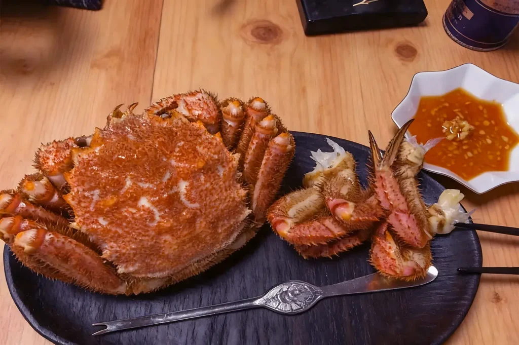 Whole boiled crab with legs on a black plate, chopsticks, a butter knife, and a small dish of dipping sauce on a wooden table.